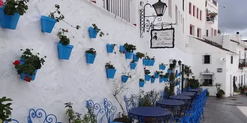 Una calle de Mijas Pueblo con floreros de geranios colgados en el muro y unas mesas para tomarte algo debajo.